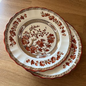 May include: Two white ceramic plates with a red and gold floral pattern. The plates have a scalloped edge and a decorative border.