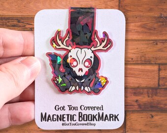 May include: A magnetic bookmark featuring a stylized skull with antlers, red eyes, and colorful geometric accents. The bookmark is on a white card with the text "Got You Covered MAGNETIC BOOKMARK" and the shop's Instagram handle.