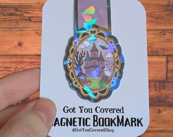 May include: A magnetic bookmark with a Halloween theme. The bookmark features a spooky haunted house, a ghost, and a spider web design. The text "Got You Covered MAGNETIC BOOKMARK" is printed at the bottom. The bookmark has a holographic finish.