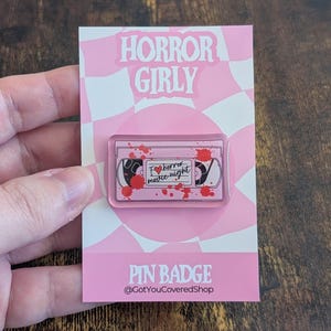 May include: A pink pin badge with the text "HORROR GIRLY" and "PIN BADGE". The pin features a cassette tape design with the words "I heart horror movie night" and red blood splatter details.