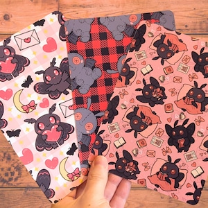 Peut inclure: Trois feuilles de tissu décoratives avec des motifs répétitifs. L'une présente un motif à carreaux roses et blancs avec des illustrations de papillons de nuit tenant des cœurs. Une autre a un motif à carreaux rouges et noirs avec des figures de poupées. La troisième a un fond rose avec des illustrations de papillons de nuit lisant des livres.