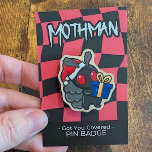 May include: A Mothman pin badge on a red and black chequered display card. The pin features a black creature with red eyes wearing a red and white Santa hat, holding a blue gift box with a yellow ribbon. The card text reads MOTHMAN and PIN BADGE.