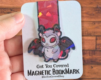 May include: A magnetic bookmark featuring a cartoon demon-like creature with red eyes, horns, and wings. The bookmark has a red, glittery top and the text "Got You Covered MAGNETIC BOOKMARK" on a white card.