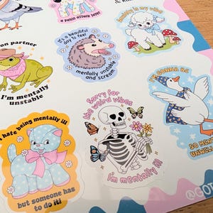 May include: A sticker sheet featuring various illustrated characters including a frog in a cowboy hat, an opossum, a lamb, a goose in a wizard hat, a blue polka-dot cat, and a skeleton holding flowers, all accompanied by humorous text.
