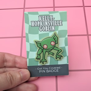 May include: A green goblin pin badge featuring large pink eyes and pointed ears, mounted on a light green and white checkered display card. The card displays the text KELLY HOPKINSVILLE GOBLIN and Got You Covered PIN BADGE in black lettering.