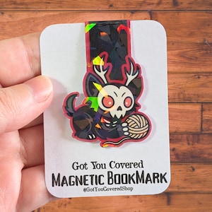 May include: A magnetic bookmark with a black cat-like creature with a skull face, antlers, and a ball of yarn. The bookmark has a geometric design with red, yellow, and green accents. Text on the card reads "Got You Covered MAGNETIC BOOKMARK".