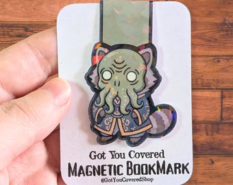 May include: A magnetic bookmark featuring a cartoon creature with tentacles, wearing a blue coat. The bookmark has the text "Got You Covered MAGNETIC BOOKMARK" and the shop's Instagram handle. The bookmark is on a white card.