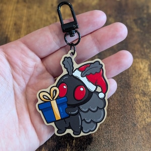 May include: A black moth character keyring with large red eyes, wearing a red and white Santa hat, and holding a blue gift box with a yellow ribbon. The charm is attached to a black metal lobster clasp hardware for easy attachment.