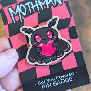 May include: A Mothman pin badge featuring a black creature with large pink eyes holding a bright pink heart. The pin is attached to a backing card with a pink and black checkerboard pattern, the text MOTHMAN, and Got You Covered - PIN BADGE.