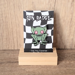 May include: A pin badge featuring a green goblin with pink eyes and a wide grin, set against a black and white checkered background. The words "PIN BADGE" are at the top. The pin is displayed on a wooden block.