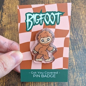 May include: A brown Bigfoot character pin badge on a backing card with a pink and brown checkered pattern. The card features the text BIGFOOT at the top and Got You Covered PIN BADGE at the bottom. The pin is a clear acrylic shape of the creature.