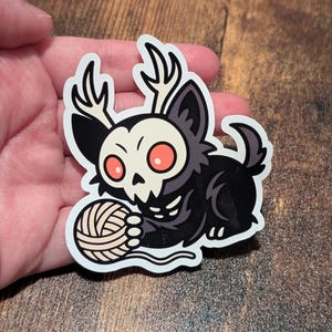 May include: A die-cut sticker depicting a black creature with a skull-like face, antlers, and red eyes, holding a ball of yarn. The sticker has a white border and is approximately 7.6 cm wide.