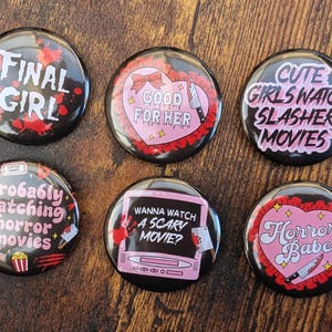 May include: Six black pin badges with horror movie themes. Each badge features text such as "FINAL GIRL" and "CUTE GIRLS WATCH SLASHER MOVIES" in white and pink, with decorative elements including hearts and knives.