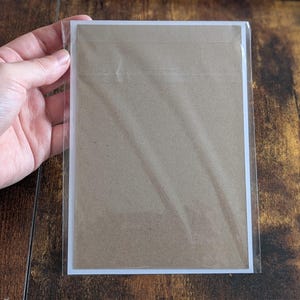 May include: A rectangular piece of brown kraft paper cardstock enclosed in a clear plastic protective sleeve. The cardstock features a plain, solid brown surface with a white border frame, presented as a flat, vertical rectangular item.