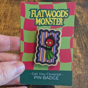 Könnte beinhalten: Ein rechteckiger Ansteck-Pin mit einer Illustration des Flatwoods-Monsters auf einer gewellten, grün-rot karierten Trägerkarte. Der Pin zeigt eine rotköpfige Kreatur in einem grünen Kleid vor einem lila-orange gestreiften Hintergrund mit Wellenrand.