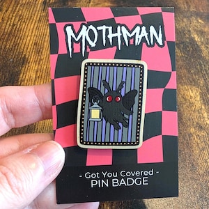 May include: A rectangular wooden pin badge featuring a black Mothman character with red eyes holding a yellow lantern. The pin is set against a purple and grey striped background with a dotted border, mounted on a pink and black chequered card.