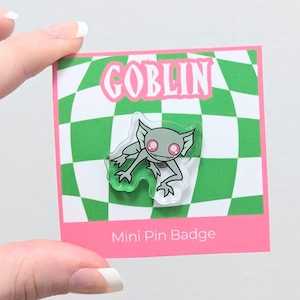 May include: A mini pin badge featuring a cartoon goblin design. The goblin is gray with pink eyes and green outlines. The pin is on a pink and green checkered background with the word "GOBLIN" in pink letters.