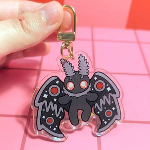 May include: A clear acrylic keychain featuring a stylized moth character with a dark gray body, fluffy light gray chest, and large orange eyes. The moth has dark gray wings decorated with red circular spots and white star patterns, attached to a gold clasp.
