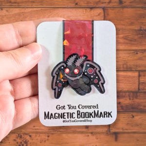May include: A magnetic bookmark featuring a cartoon moth design with a black body, gray wings, and red accents. The bookmark has the text "Got You Covered MAGNETIC BOOKMARK" and the shop's Instagram handle.