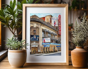 May include: A framed watercolor painting of a theater building with a red marquee that reads "The NoteBook" and "Schoenfeld". The building has a red brick facade and a white awning. The painting is set against a blue sky with white clouds.