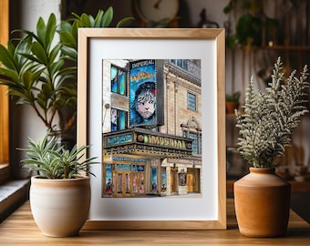 May include: A framed watercolor print of the Imperial Theatre, showcasing the marquee for "Les Misérables." The artwork captures the building's facade with detailed architectural elements, including signage and windows. The print is displayed on a wooden table.