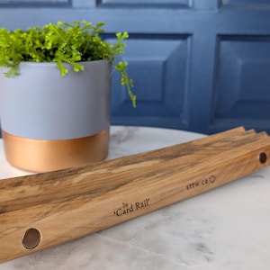 Puede incluir: Un riel para tarjetas rectangular de madera con acabado de veta natural, que presenta dos imanes circulares incrustados en la parte frontal. El texto 'The Card Rail' y 'sttw.ca' con un pequeño icono de hoja de arce están impresos en la madera.
