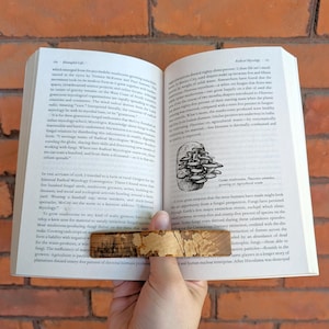 May include: A wooden bookmark with a natural wood grain pattern is placed in an open book. The book's pages are visible, with text and illustrations. The bookmark is held in place, suggesting a reading session or a display of the bookmark's function.