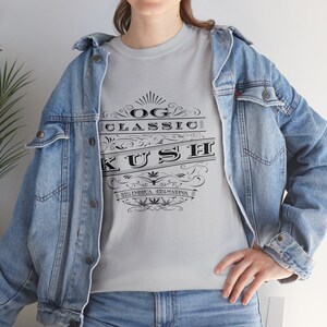 May include: A gray t-shirt with a black graphic design that says "OG Classic Kush" with a stylized font and a cannabis leaf design. The graphic also includes the text "50% Indica 50% Sativa".