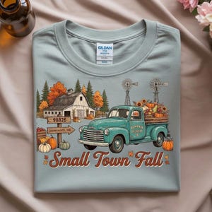 May include: A light blue t-shirt featuring a graphic of a teal pickup truck, a white barn, windmills, and pumpkins. The text reads Pumpkin Delivery Co. and Small Town Fall, with a signpost marked 98826 Leavenworth, WA. The shirt has a Gildan label.