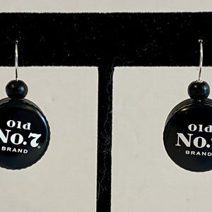 Może przedstawiać: Para czarnych kolczyków z srebrnymi haczykami. Każdy kolczyk ma czarną zakrętkę od butelki z napisem "Old No. 7 Brand" nadrukowanym na biało.