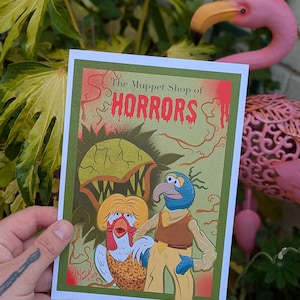 May include: A print with the text "The Muppet Shop of HORRORS" and a cartoon illustration. The artwork shows two Muppet characters in front of a large, green, plant-like creature. The print has a green border and is set against a background of foliage.
