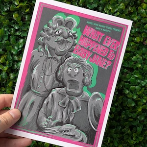 Op de afbeelding: Een print met Statler en Waldorf van The Muppets, in een parodie op de film "What Ever Happened to Baby Jane?" Het kunstwerk is in grijstinten, met roze en groene accenten. De tekst "Muppet Movie Mashup Studios Presents" staat bovenaan.