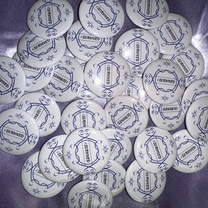May include: A collection of round, white, and blue pins with the text "I Survived" in a decorative font. The pins are arranged in a random pattern on a purple surface.