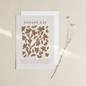 Op de afbeelding: Een witte kaart met een gouden bloemenpatroon en de tekst "Romeinen 8:28" en "Want wij weten, dat voor hen die God liefhebben, alle dingen medewerken ten goede, voor hen die naar zijn voornemen geroepen zijn."