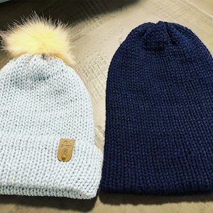 Puede incluir: Dos gorros de punto, uno azul claro con un pompón amarillo y otro azul oscuro. Ambos gorros tienen un pequeño parche de cuero con un logotipo.