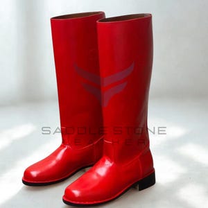 Puede incluir: Un par de botas altas de cuero rojo brillante. Estas botas tienen un acabado brillante, una puntera redondeada y un tacón bajo negro. El nombre de la marca "SADDLE STONE LEATHER" está impreso en el lateral.
