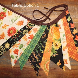 Könnte beinhalten: Eine Stoffwimpelkette mit der Aufschrift Fabric Option 1, bestehend aus dreieckigen Wimpeln mit herbstlichen Mustern wie Kürbissen, Sonnenblumen, Herbstblättern und Karomustern in Orange, Grün, Schwarz und Creme, verbunden durch ein braunes Band.