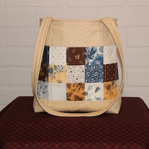 May include: A beige patchwork tote bag with blue, brown, and yellow floral patterns. The bag has two long straps and a quilted design.