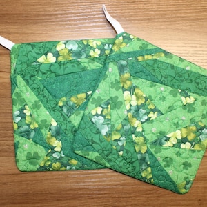 Puede incluir: Dos agarraderas verdes acolchadas con lazos blancos para colgar. Las agarraderas presentan un diseño de patchwork con varios tonos de tela verde y un patrón de tréboles y detalles florales. Las agarraderas miden aproximadamente 20 cm cuadrados.