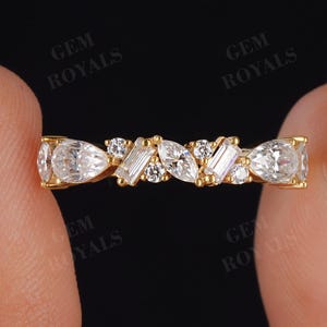 May include: A gold ring with a series of clear, sparkling gemstones. The ring features a mix of shapes, including pear, baguette, and round cut stones. The ring is held between two fingers against a black background.