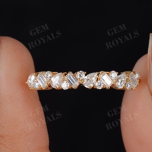 May include: A gold band ring featuring a series of alternating clear gemstones in various shapes, including round, pear, and baguette cuts. The ring is held between two fingers against a dark background, showcasing the ring's sparkle and design.
