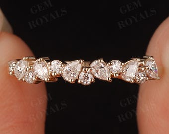 May include: A gold ring with a series of clear, sparkling gemstones in various shapes, including pear and round cuts. The ring is held between two fingers against a dark background, showcasing the ring's intricate design.