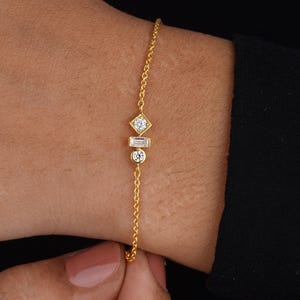 Könnte beinhalten: Ein zartes Armband aus einer goldenen Kette mit drei kleinen Diamanten. Die Diamanten sind in quadratischer, Baguette- und runder Form gefasst.