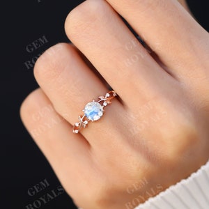 Puede incluir: Un delicado anillo de oro rosa con una piedra de luna redonda en el centro y un diseño floral. El anillo está engastado con diamantes más pequeños en la banda.