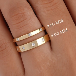 May include: Two gold rings with small clear stones set into the band. The rings are stacked on a finger. The top ring is 2.50 mm wide and the bottom ring is 4.00 mm wide.