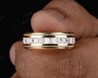 May include: A gold band ring with a row of square-cut, clear gemstones. The ring has a detailed design with a textured border. The ring is held in a hand against a dark background. The ring is a piece of jewelry.