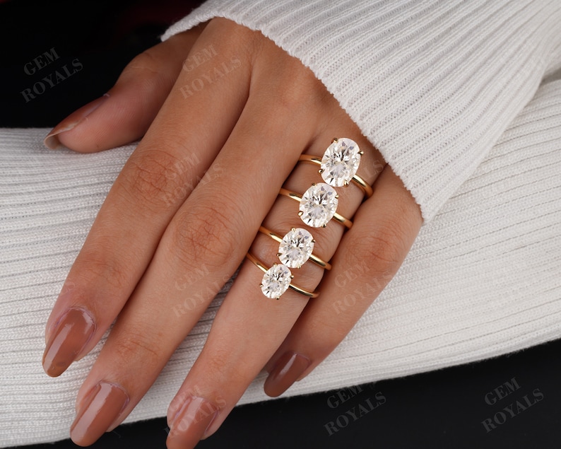 Pode incluir: Um conjunto de cinco an&eacute;is de noivado de diamantes ovais em tamanhos diferentes, engastados em bandas de ouro. Os an&eacute;is s&atilde;o exibidos em uma m&atilde;o que est&aacute; usando um su&eacute;ter branco.