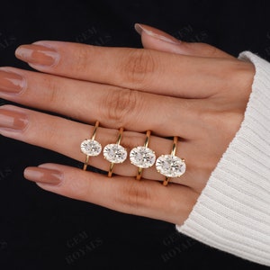 Pode incluir: Quatro an&eacute;is de noivado ovais de ouro com diamantes de tamanhos diferentes em uma m&atilde;o que est&aacute; usando um su&eacute;ter branco.