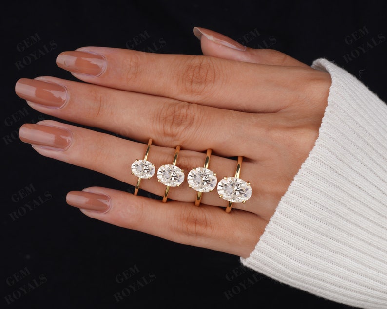 Pode incluir: Quatro an&eacute;is de noivado ovais de ouro com diamantes de tamanhos diferentes em uma m&atilde;o que est&aacute; usando um su&eacute;ter branco.