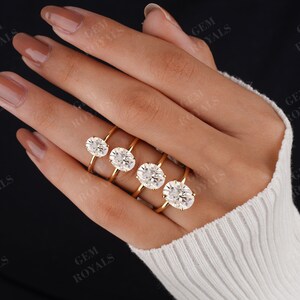 Pode incluir: Cinco alian&ccedil;as de noivado de ouro com diamantes ovais de diferentes tamanhos. As alian&ccedil;as est&atilde;o em uma m&atilde;o que est&aacute; usando um su&eacute;ter branco.